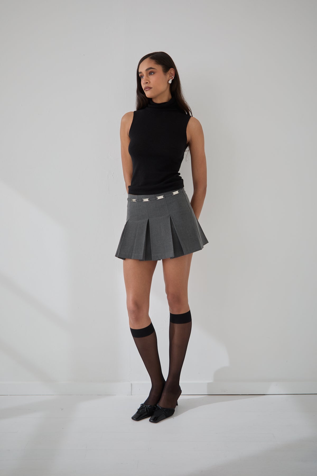 A woman standing against a white background wearing a gray mini skirt with silver tone details and a black sleeveless turtleneck.