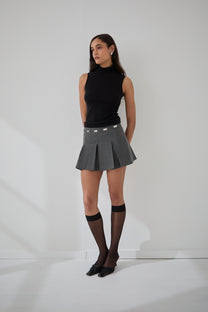 A woman standing against a white background wearing a gray mini skirt with silver tone details and a black sleeveless turtleneck.