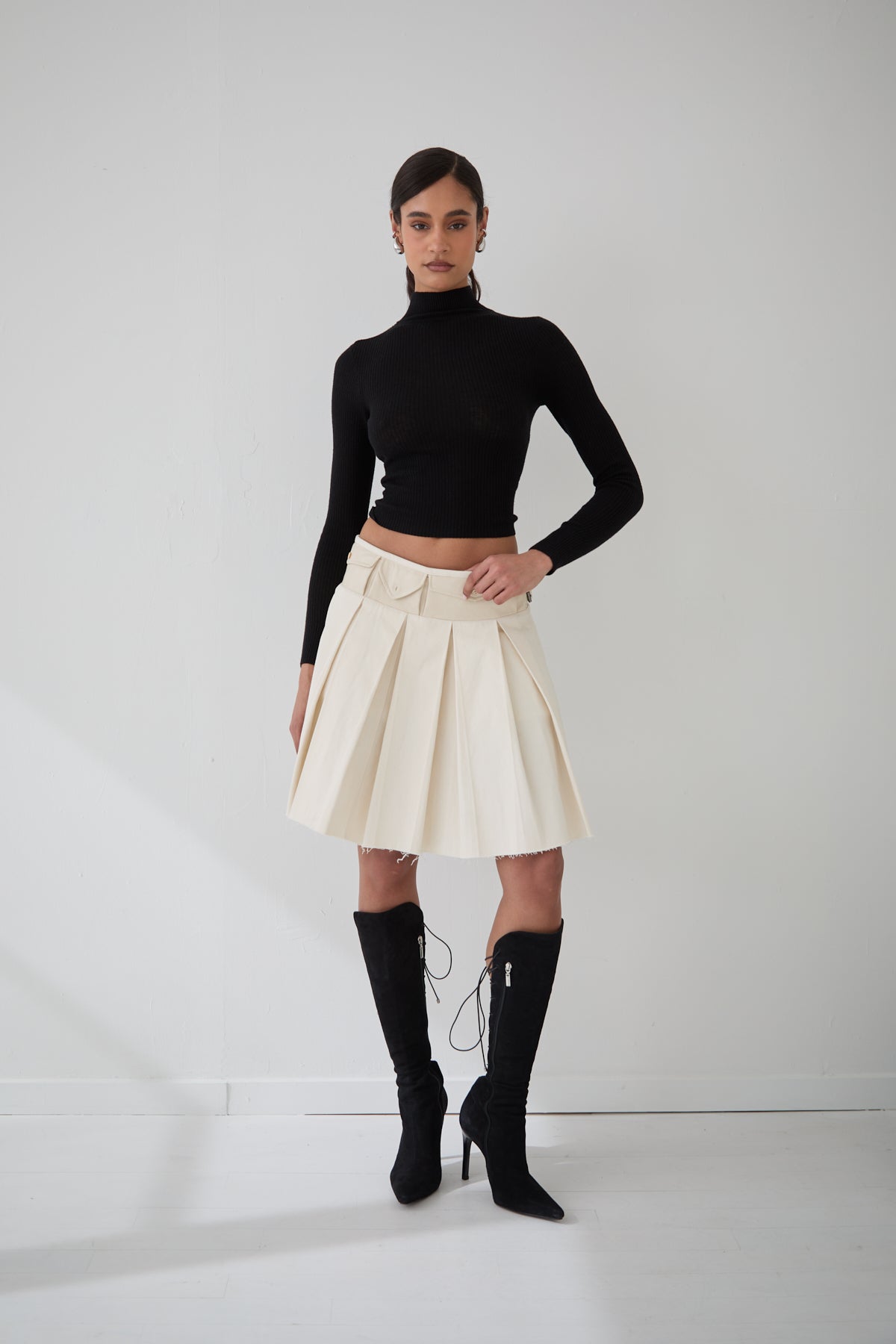 A woman wearing a black fitted long sleeve turtleneck with a rib knit pattern, paired with a cream color pleated skirt and black boots.