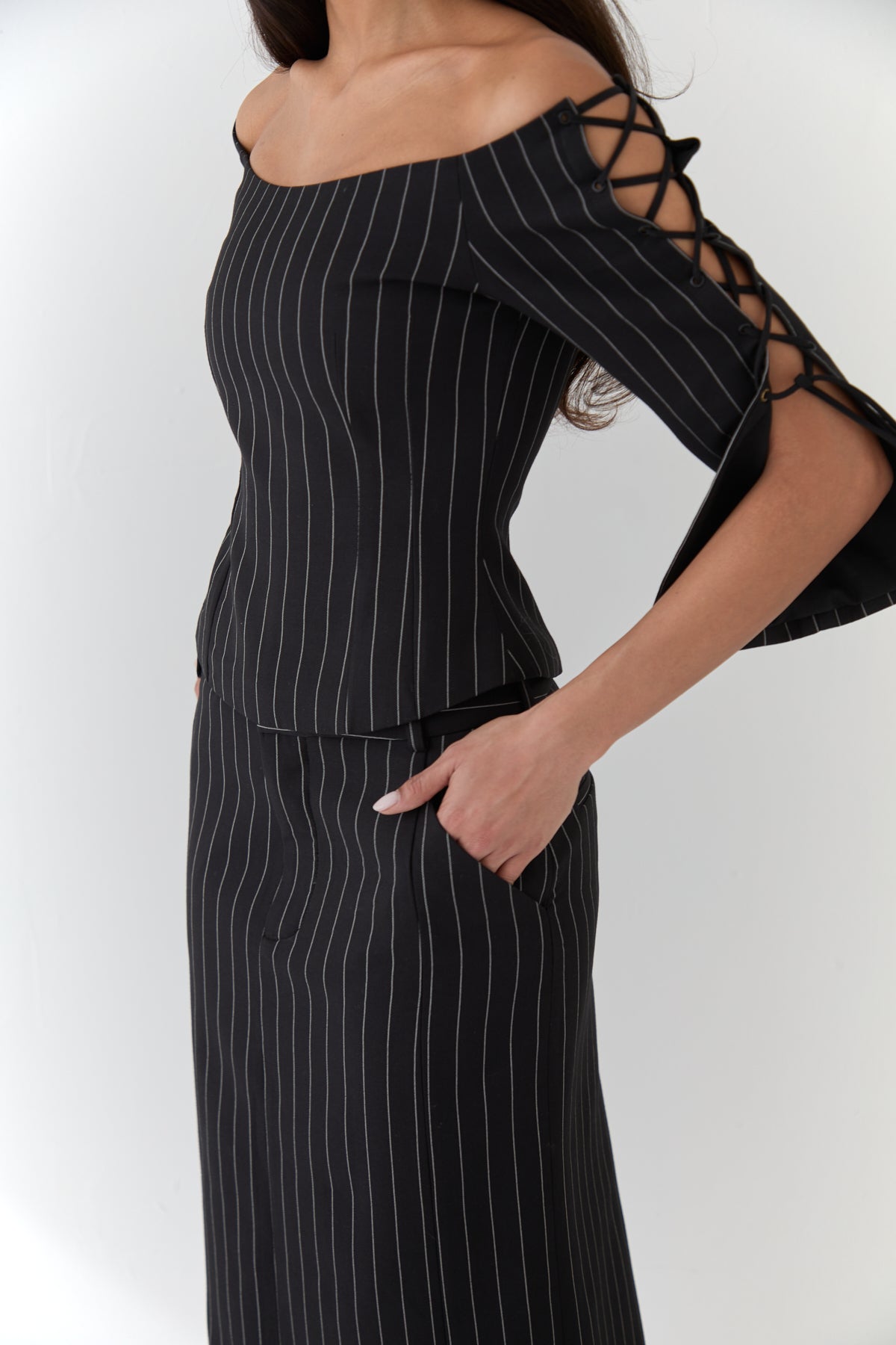A closeup of a model wearing a black pinstripe top with wide scoop neckline, lace-up 3/4 length sleeves and a matching pinstripe skirt.