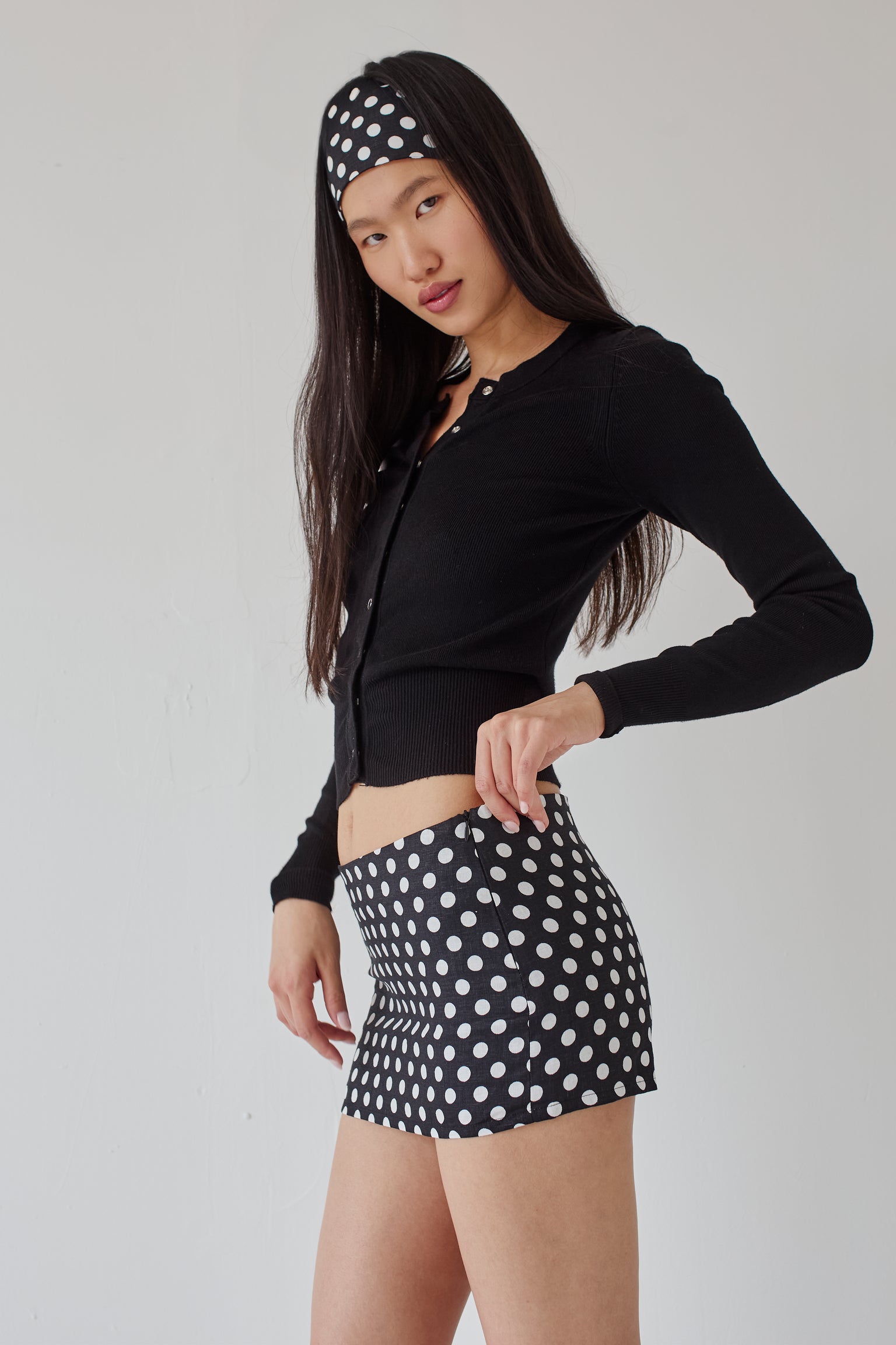 Woman wearing a black long-sleeve top, a black mini skirt with white polka dots and a matching polka dot headband on a plain background