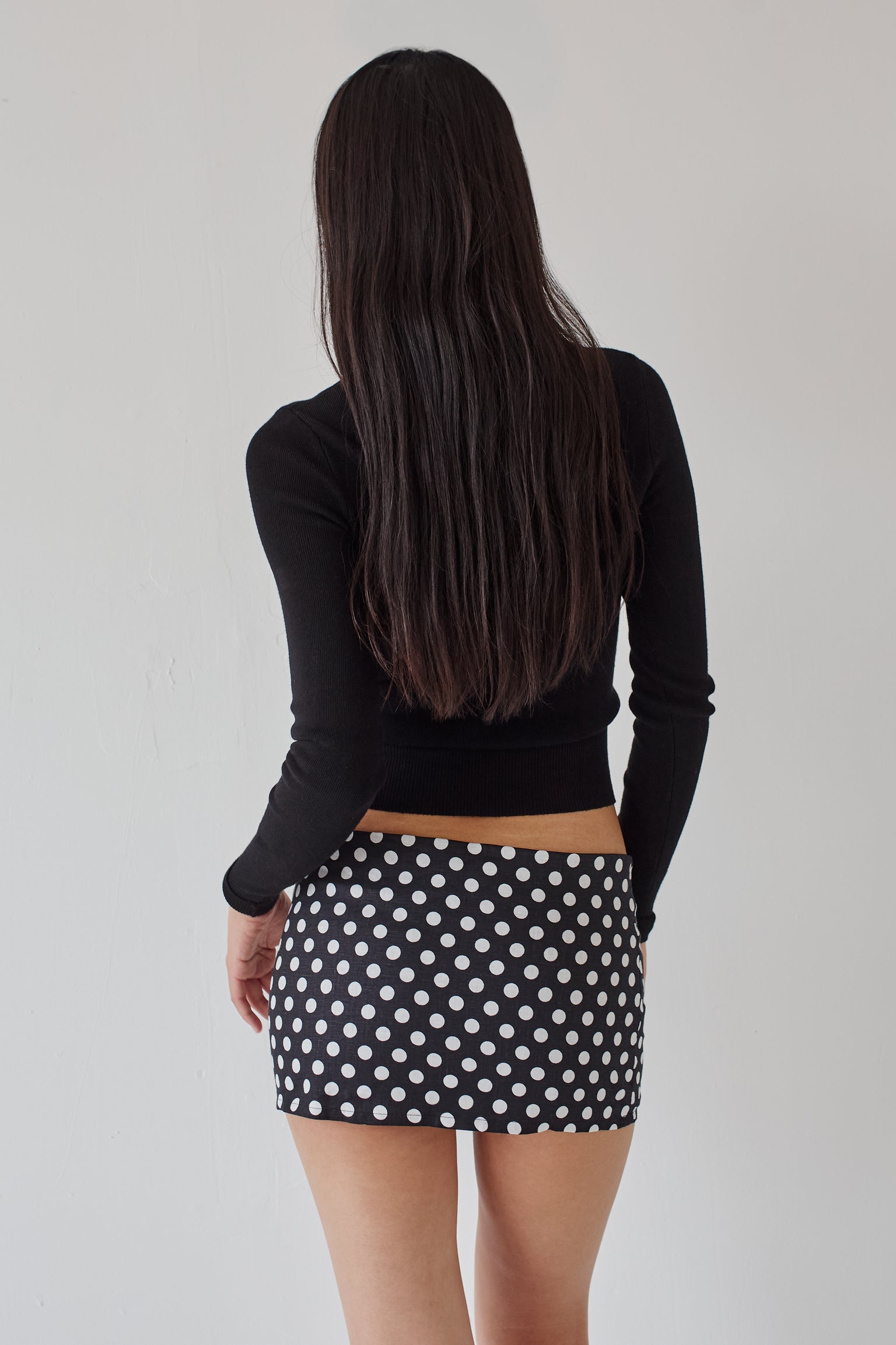 Woman wearing a black long-sleeve top and a black mini skirt with white polka dots against a white background.
