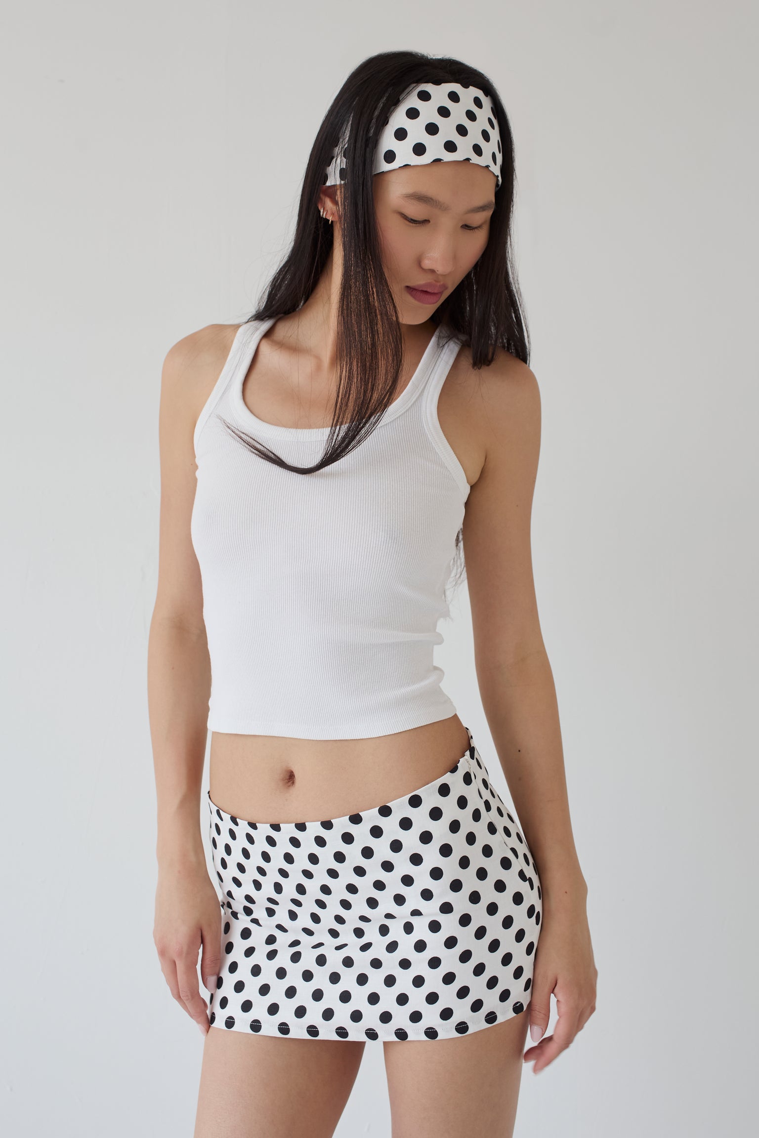 Woman wearing a white tank top and polka dot mini skirt with a matching polka dot headband on a plain background