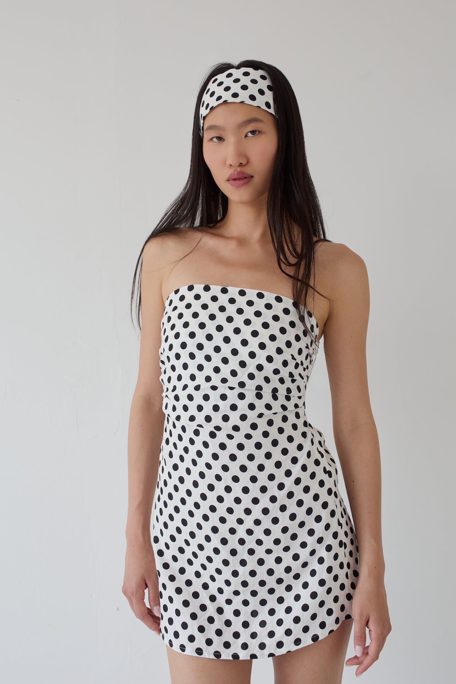 Woman wearing a white strapless dress with black polka dots and a matching polka dot headband on a plain background