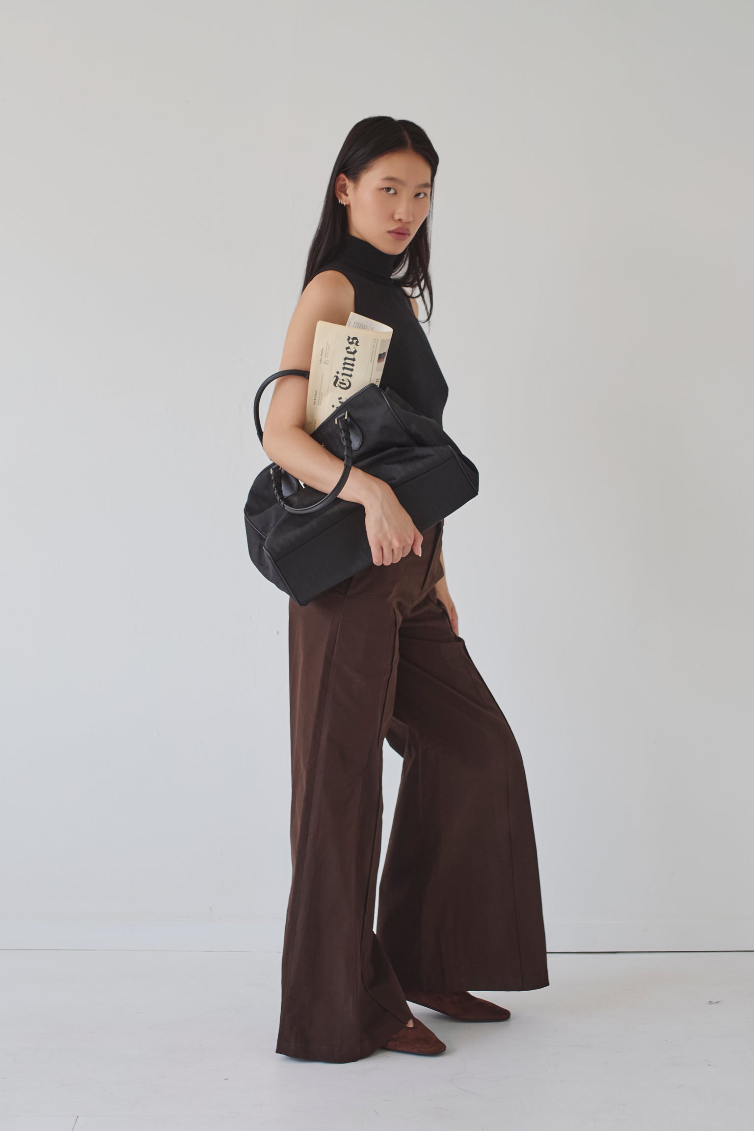Woman wearing brown wide leg pants and a sleeveless black turtleneck holding a black handbag with a newspaper in it against a white background
