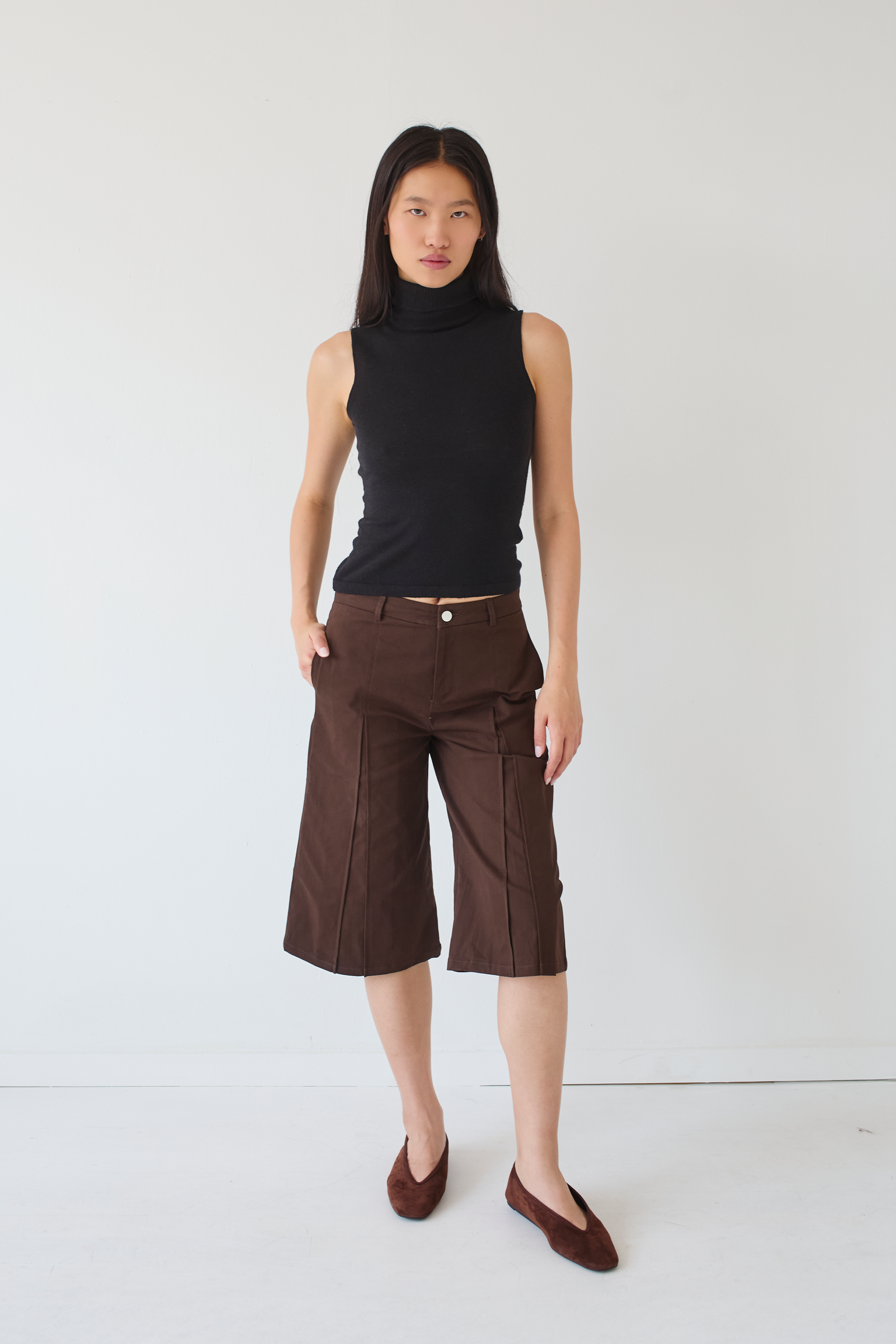 Woman wearing a black sleeveless top and brown knee length shorts against a white background