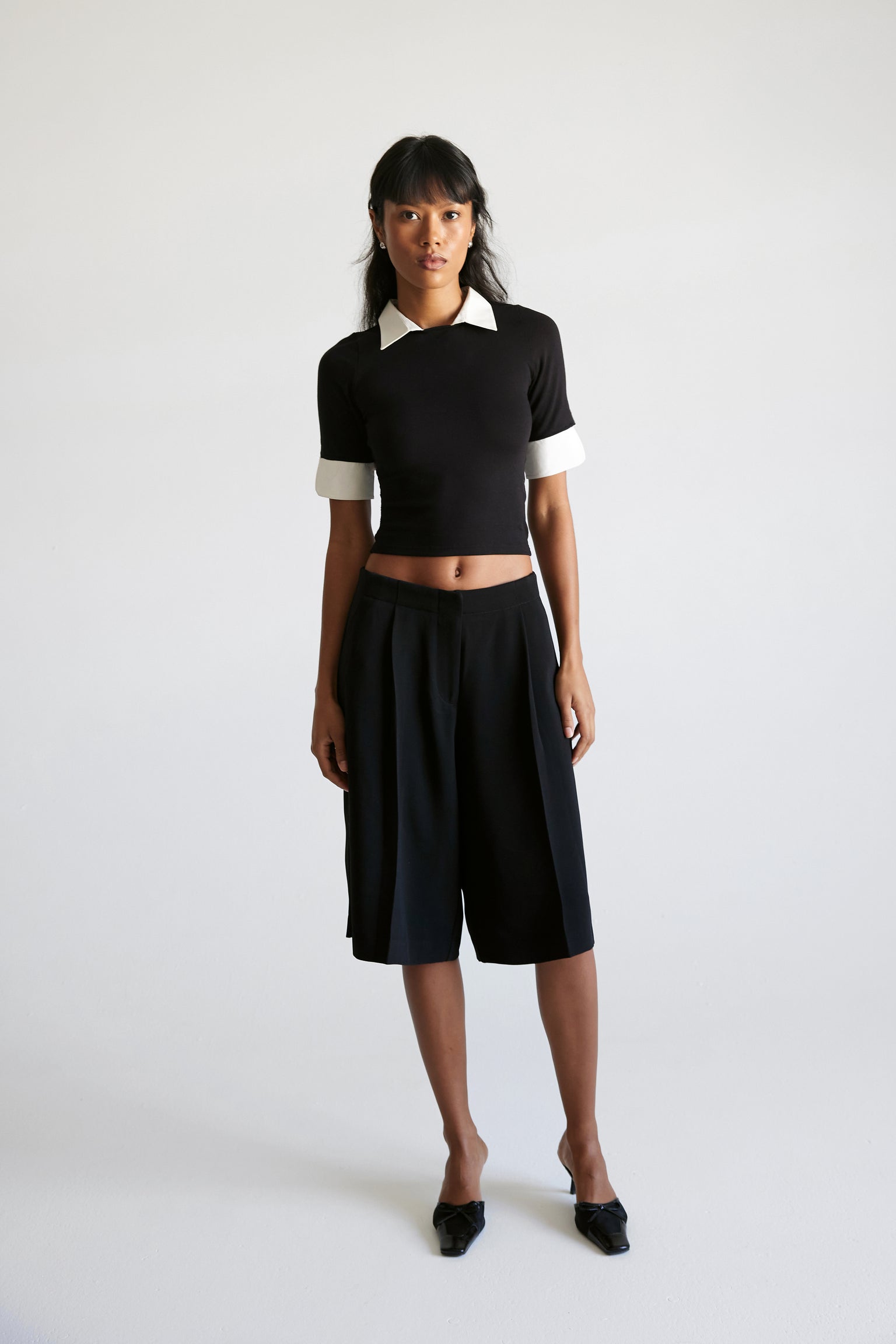 A woman standing against a white background wearing black tailored crepe shorts. She is also wearing a black crop top with contrasting collar and cuffs.