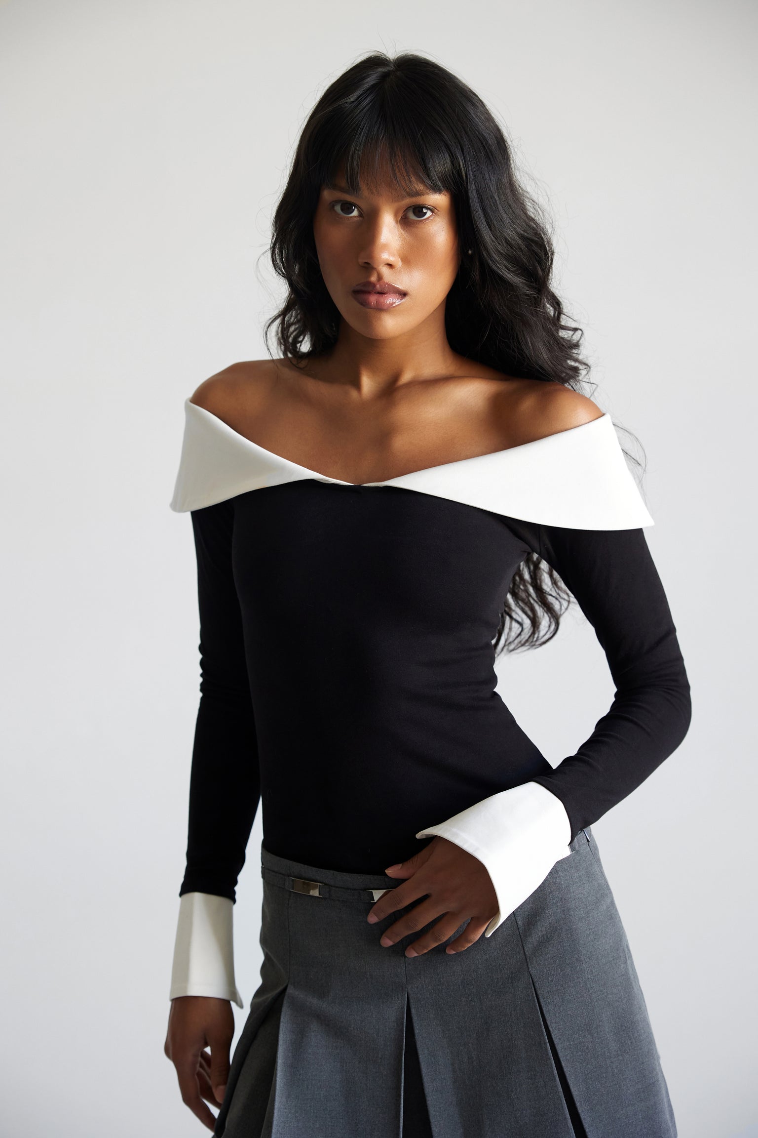 A model wearing a long sleeve off shouder top with contrasting collar and cuffs with a gray pleated mini skirt.