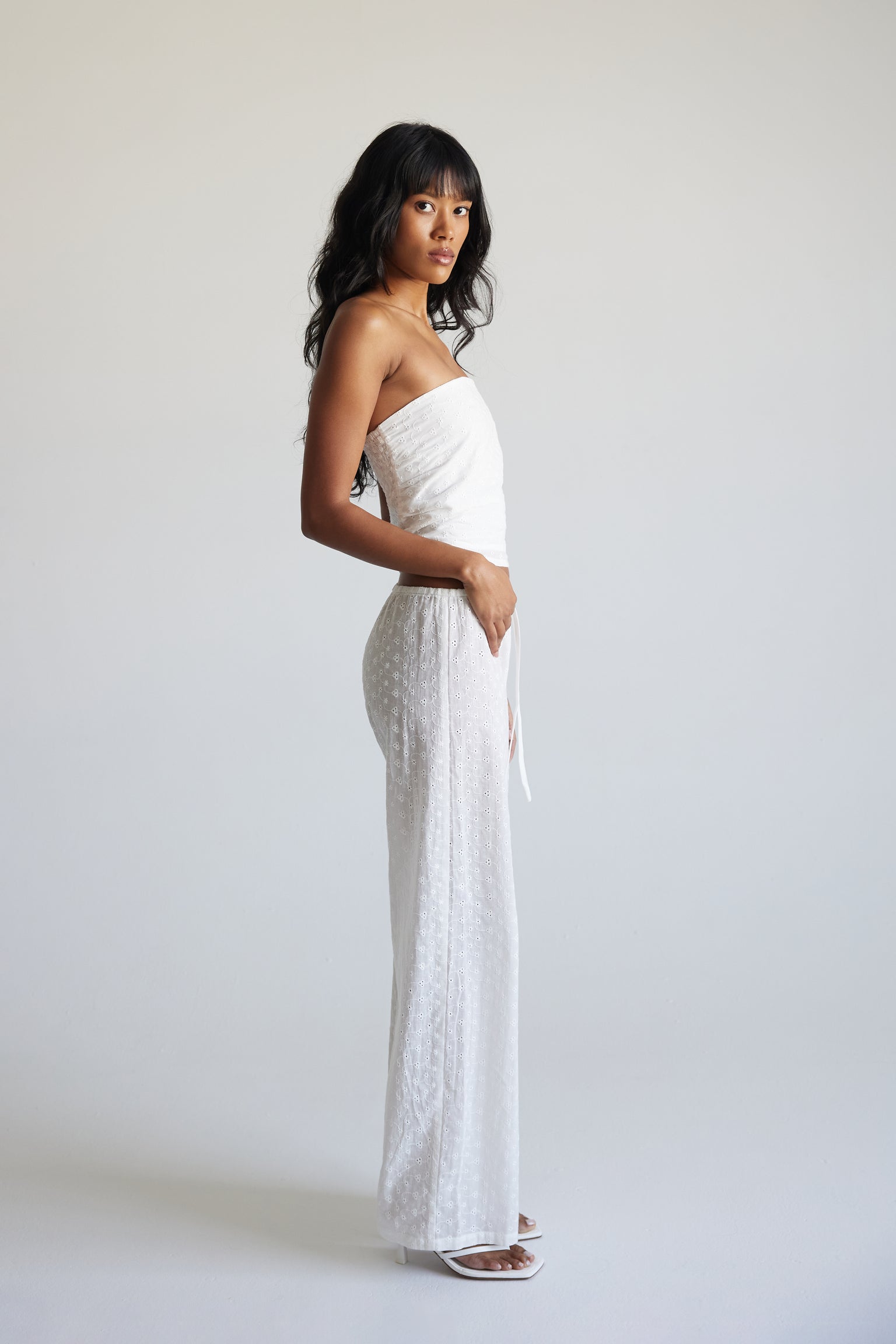 A woman wearing a white strapless top and matching white drawstring pants against a neutral background.