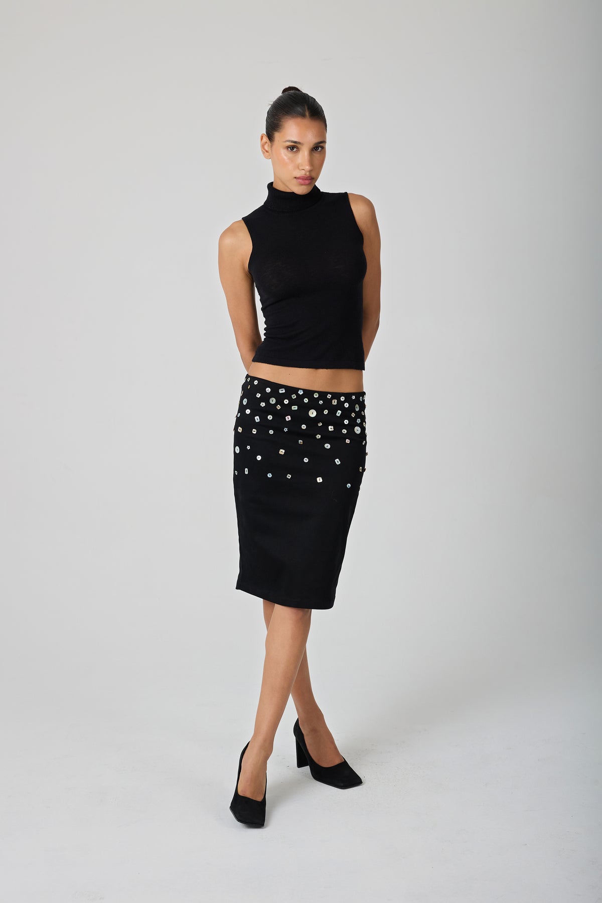 A model stands against a light gray studio background wearing a black sleeveless mock-neck top and a black knee-length skirt embellished with shell buttons.