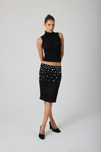 A model stands against a light gray studio background wearing a black sleeveless mock-neck top and a black knee-length skirt embellished with shell buttons.