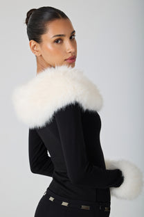 A woman wearing a black long-sleeve top with a large white fur collar and matching white fur cuffs, photographed against a light gray studio background.