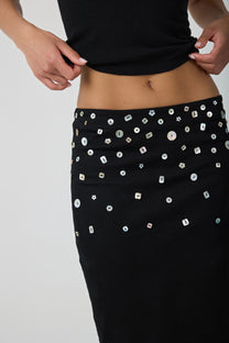 A close-up of a person wearing a black skirt embellished with shell buttons, paired with a black cropped top, against a light gray studio background.