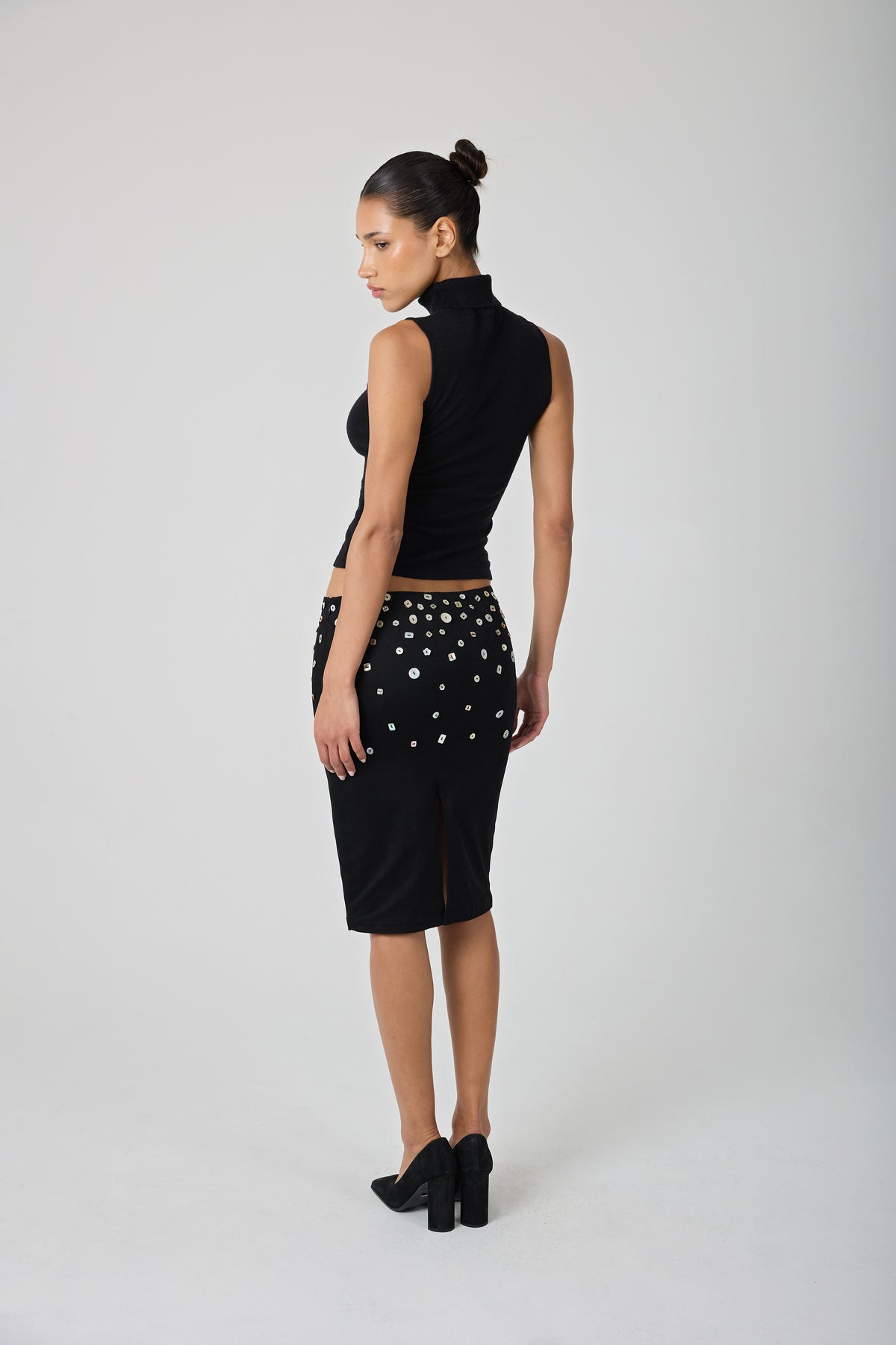 A model stands against a light gray studio background wearing a black sleeveless mock-neck top and a black knee-length skirt embellished with shell buttons.