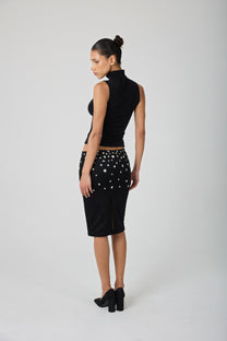 A model stands against a light gray studio background wearing a black sleeveless mock-neck top and a black knee-length skirt embellished with shell buttons.