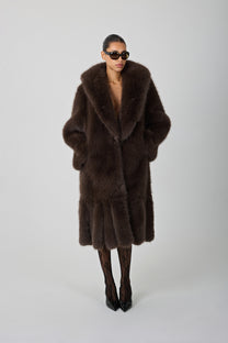 model wearing a brown fur coat and sunglasses against a blank background