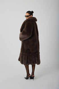 model wearing a brown fur coat and sunglasses against a blank background