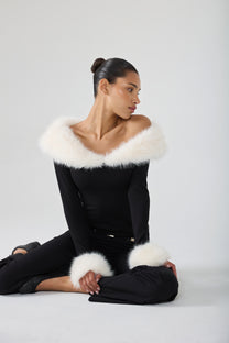 A model wearing a black off-the-shoulder long-sleeve top with a fluffy white fur collar and matching white fur cuffs, paired with black pants, posed against a light gray studio background.