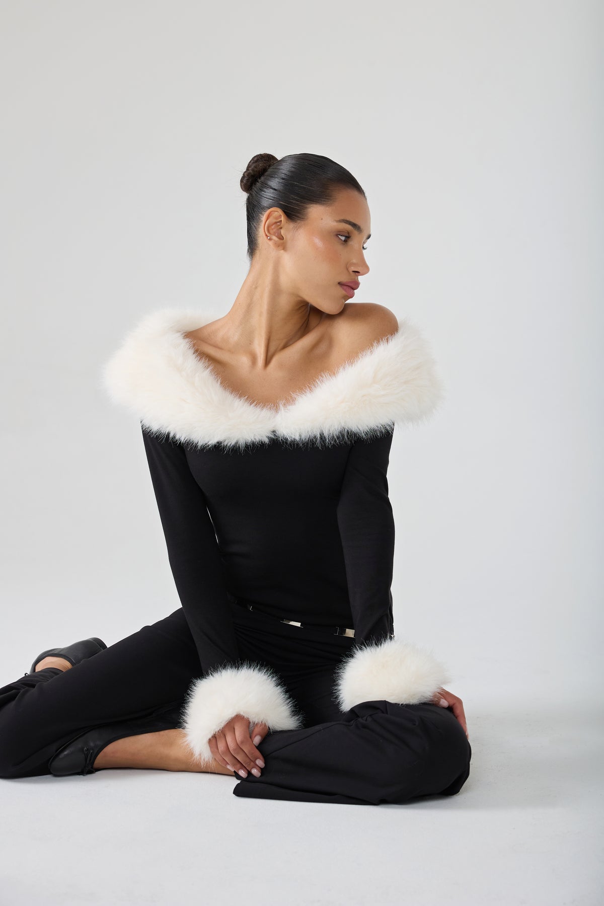 A model wearing a black off-the-shoulder long-sleeve top with a fluffy white fur collar and matching white fur cuffs, paired with black pants, posed against a light gray studio background.