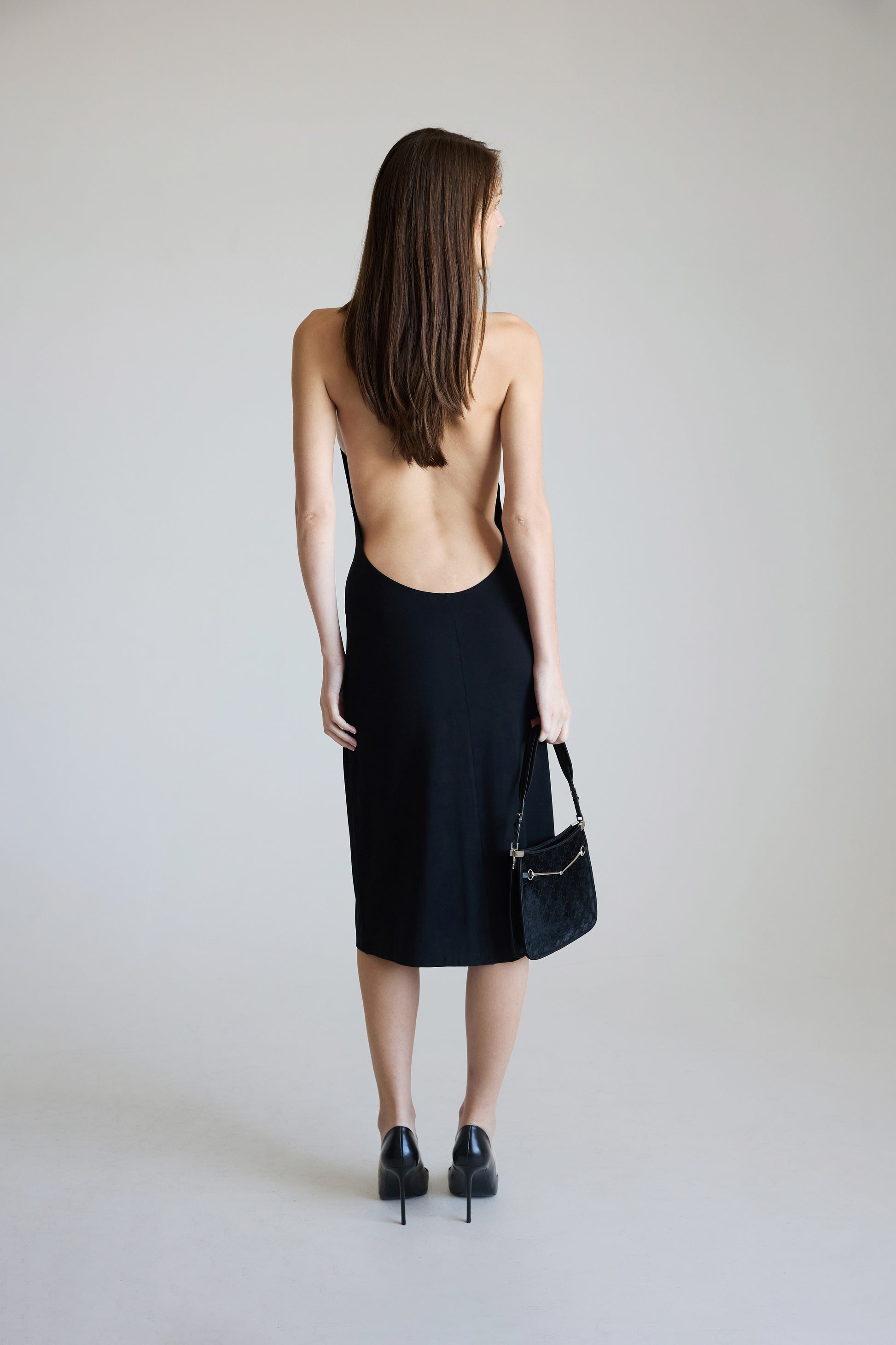 Woman wearing a black dress with an open back, holding a black handbag against a plain background