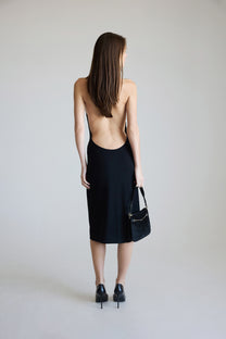 Woman wearing a black dress with an open back, holding a black handbag against a plain background