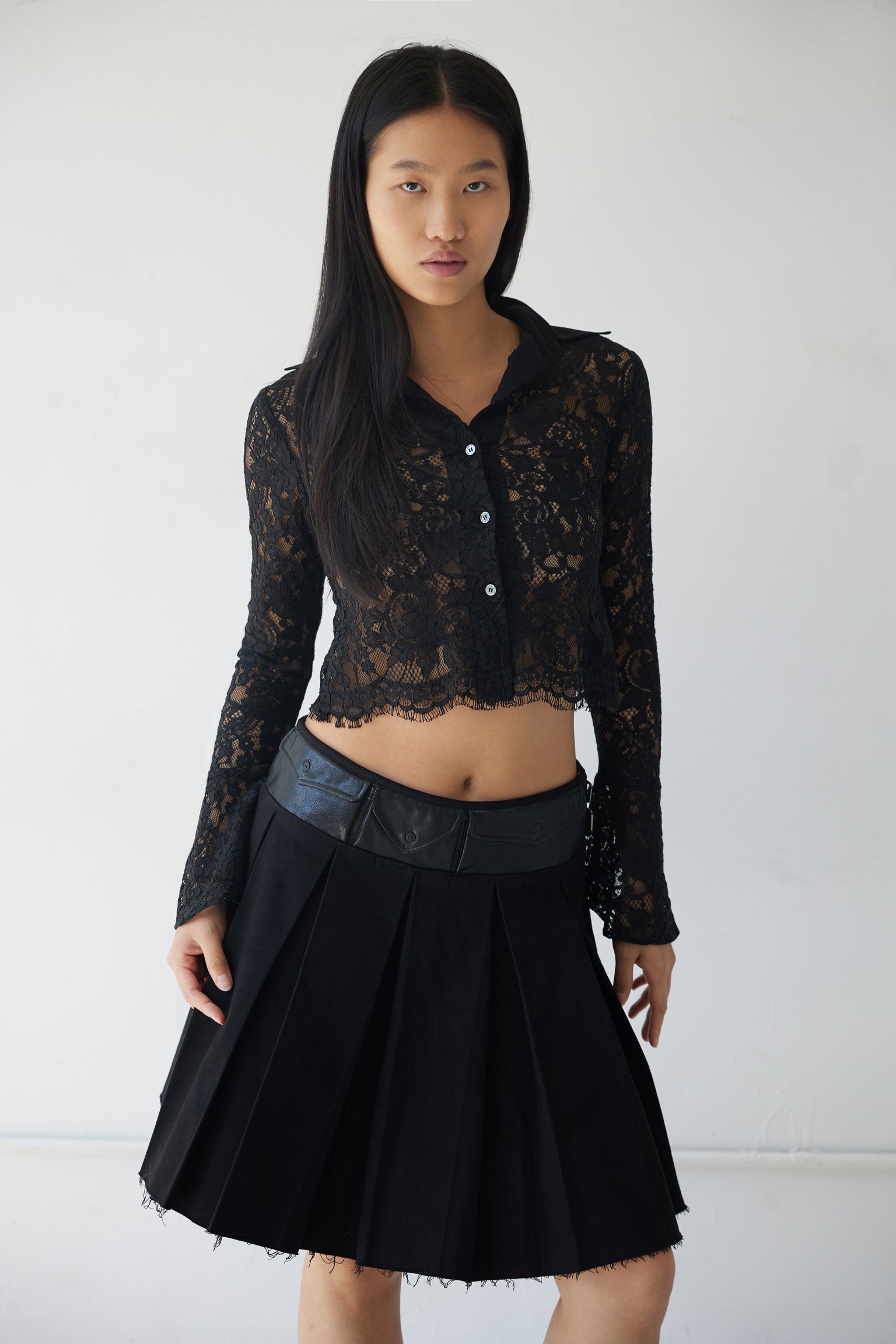 A model wearing a black box pleat midi skirt with faux leather detail pockets and a raw hem, paired with a lace long-sleeve blouse.