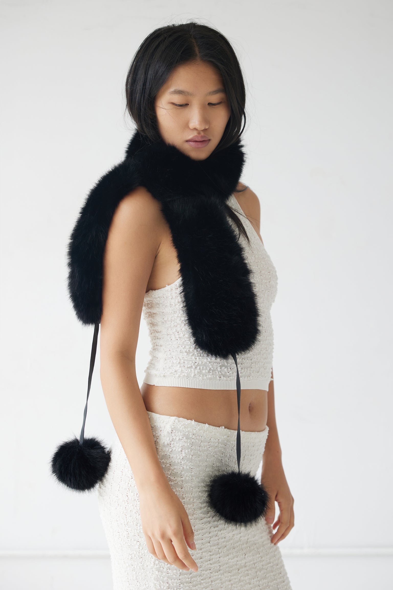 A model wearing a black faux fur scarf with pom poms, which can be styled as a neck scarf, hat, or cuffs.