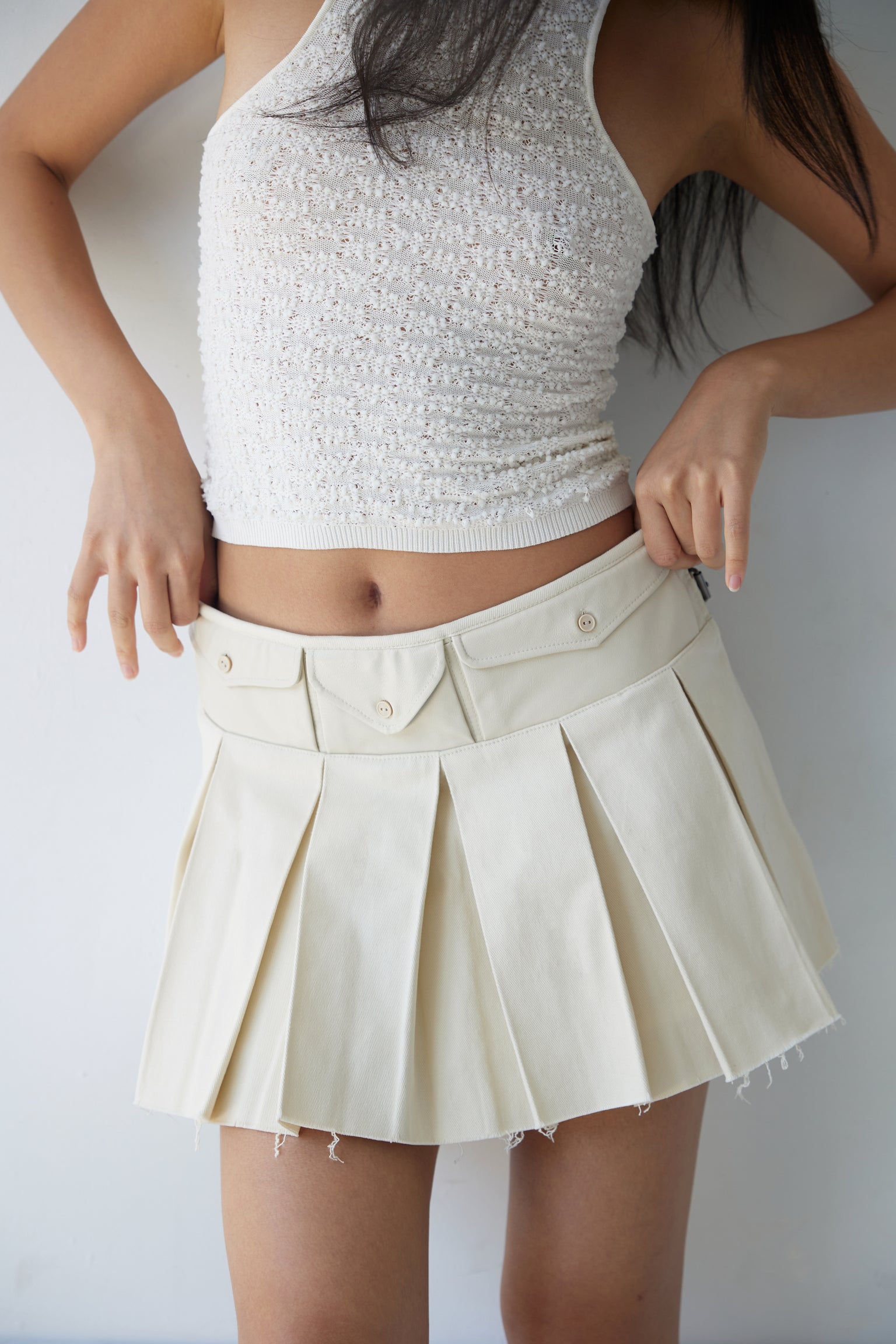 A woman wearing a beige box pleat mini skirt with faux leather pockets, paired with a white top.