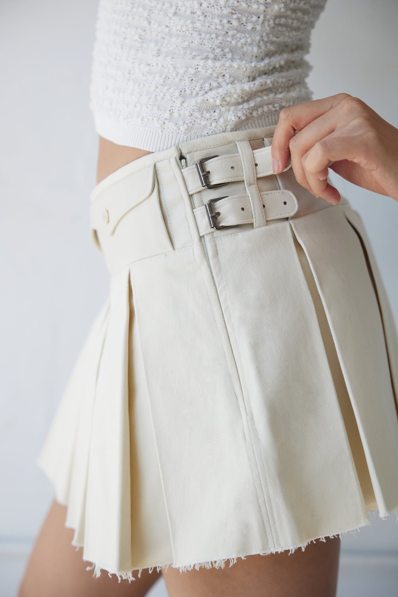 Closeup of a woman wearing a beige box pleat mini skirt with faux leather pockets and buckles, paired with a white top.