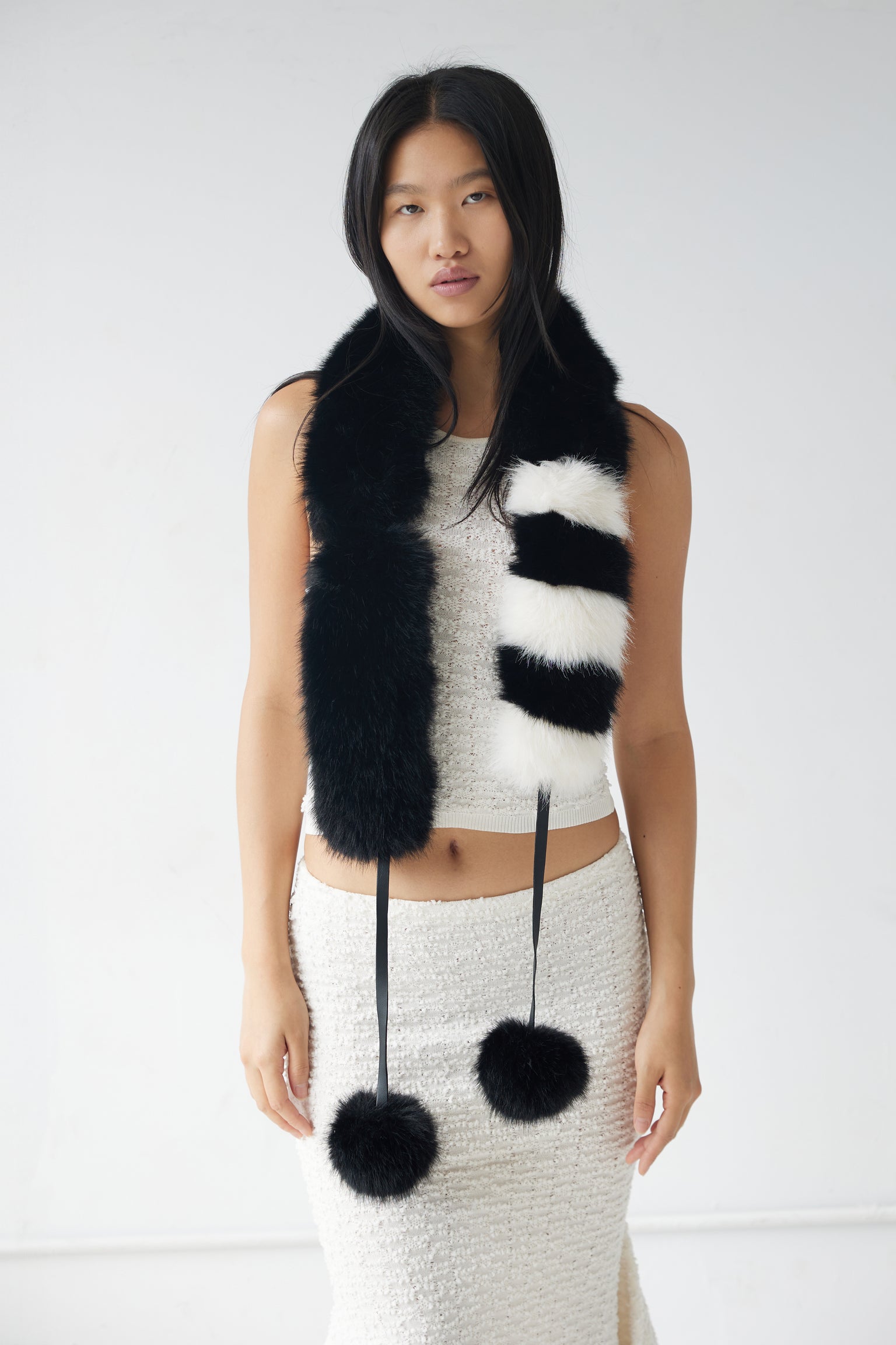 A model wearing a black and white faux fur scarf with pom poms, which can be styled as a neck scarf, hat, or cuffs.