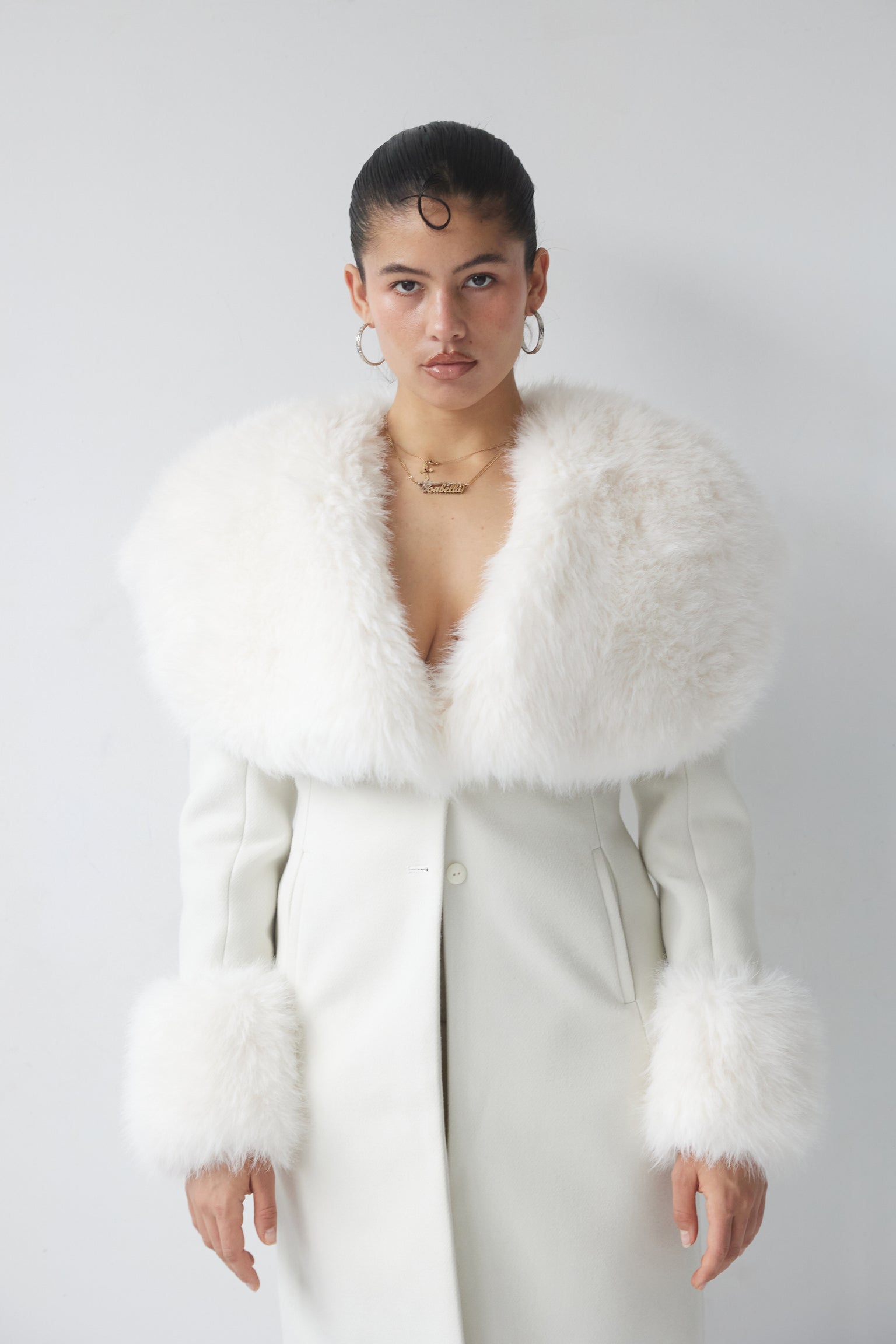 A woman wearing a white coat with a cinched waist and dramatic faux fur collar and cuffs, standing against a plain background.
