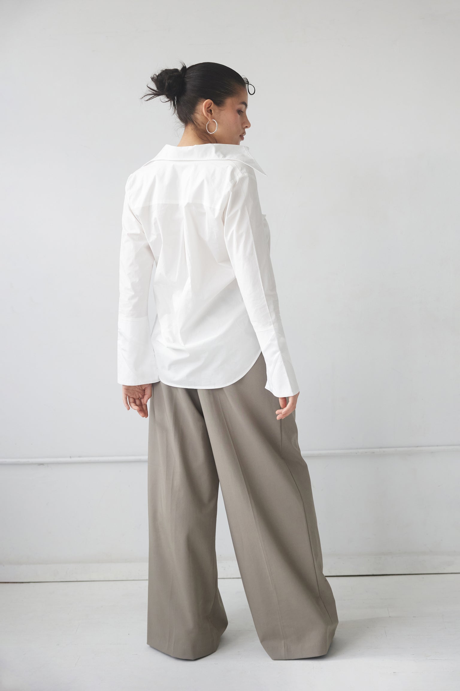 A woman wearing a white button-up shirt with an exaggerated collar and cuffs, paired with gray pants standing against a neutral background.