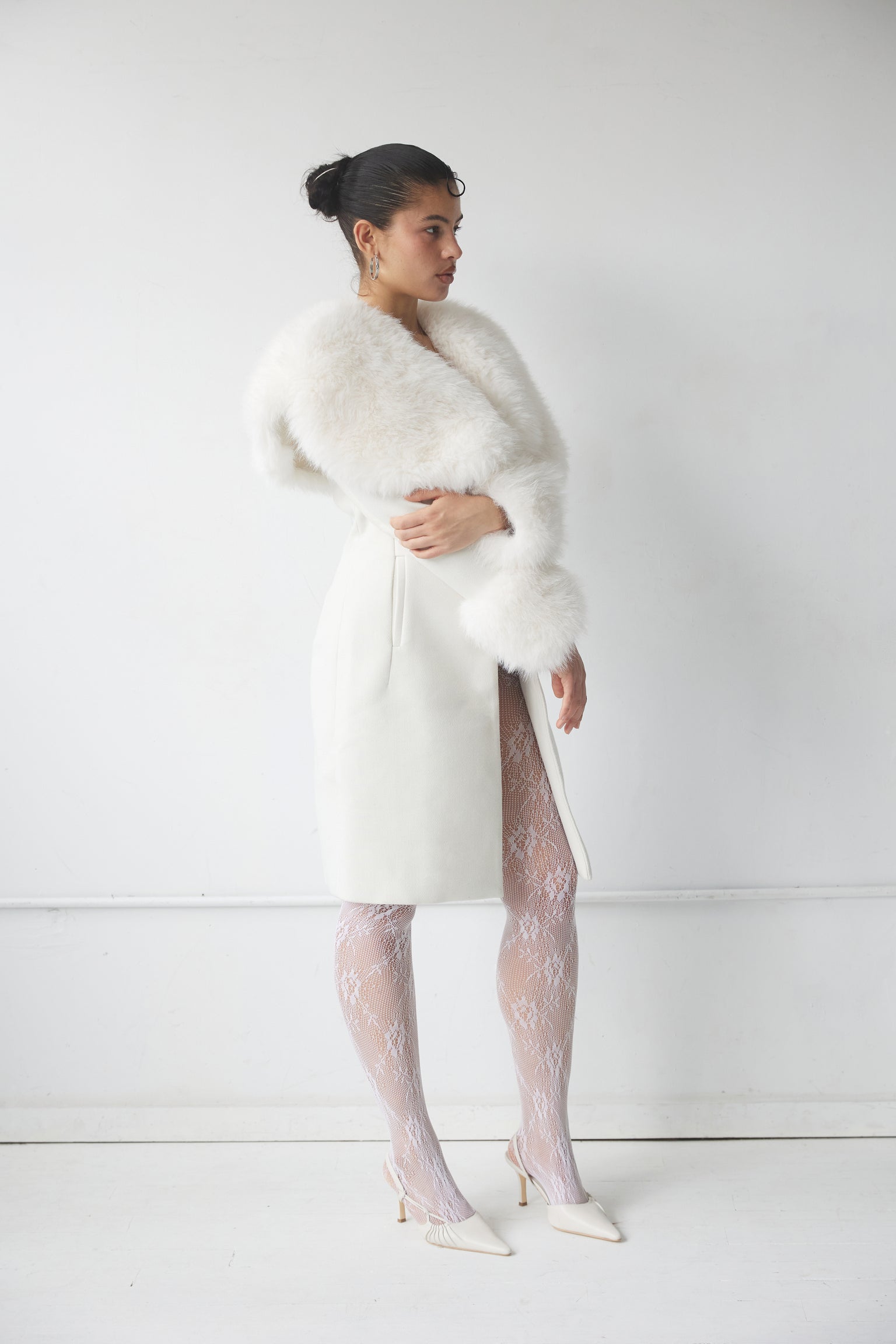 A woman wearing a white knee-length  coat with a cinched waist and dramatic faux fur collar, standing against a plain background.