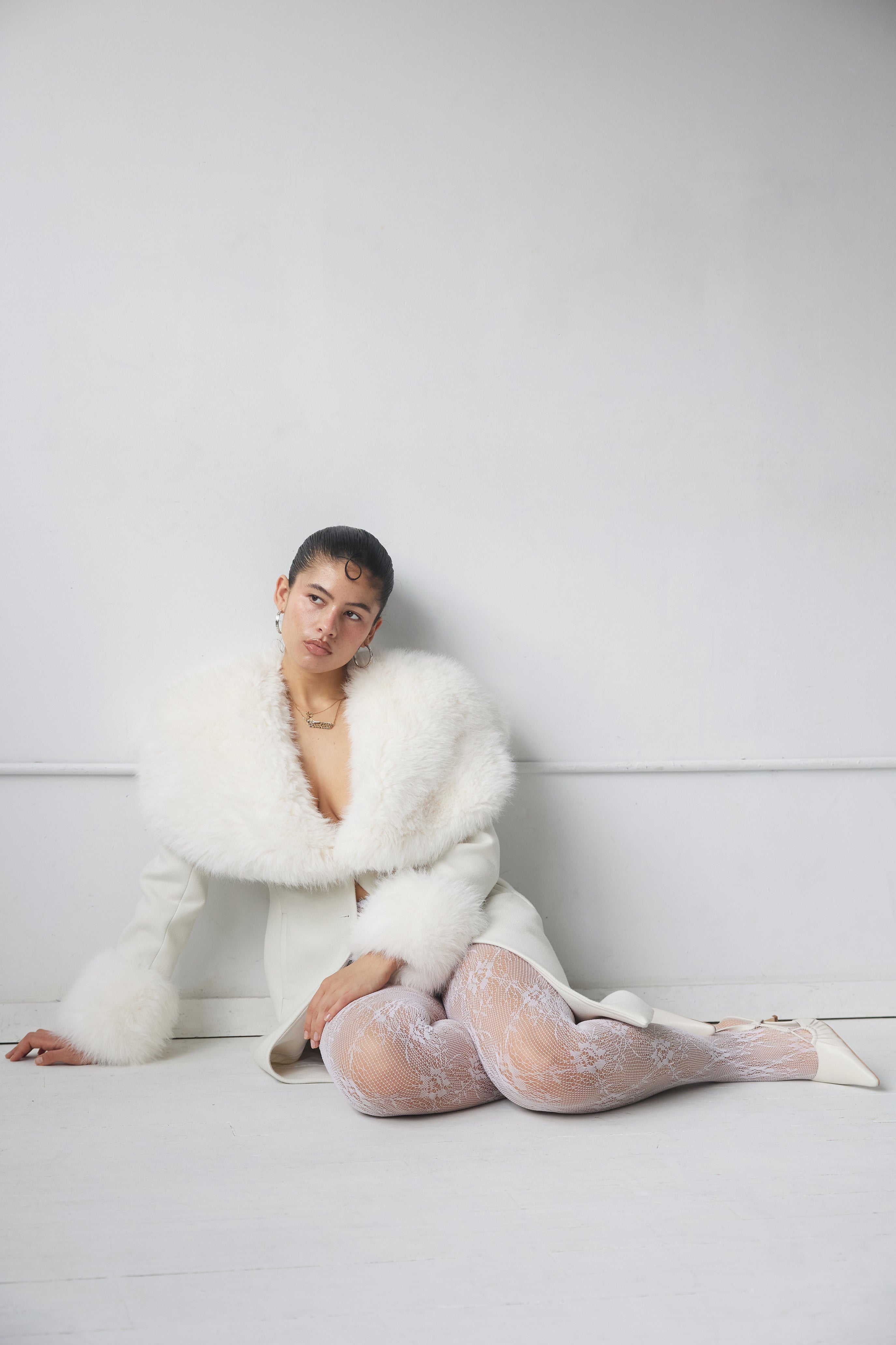 Woman wearing a white fur coat sitting on the floor against a white wall.