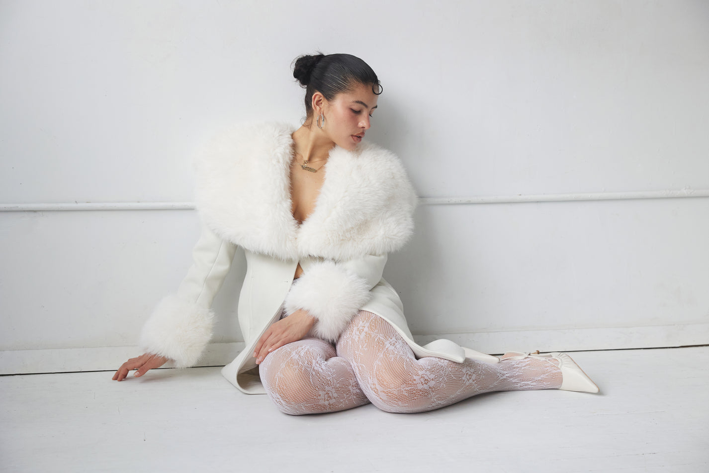Woman wearing a white fur coat and stockings sitting on a white floor.