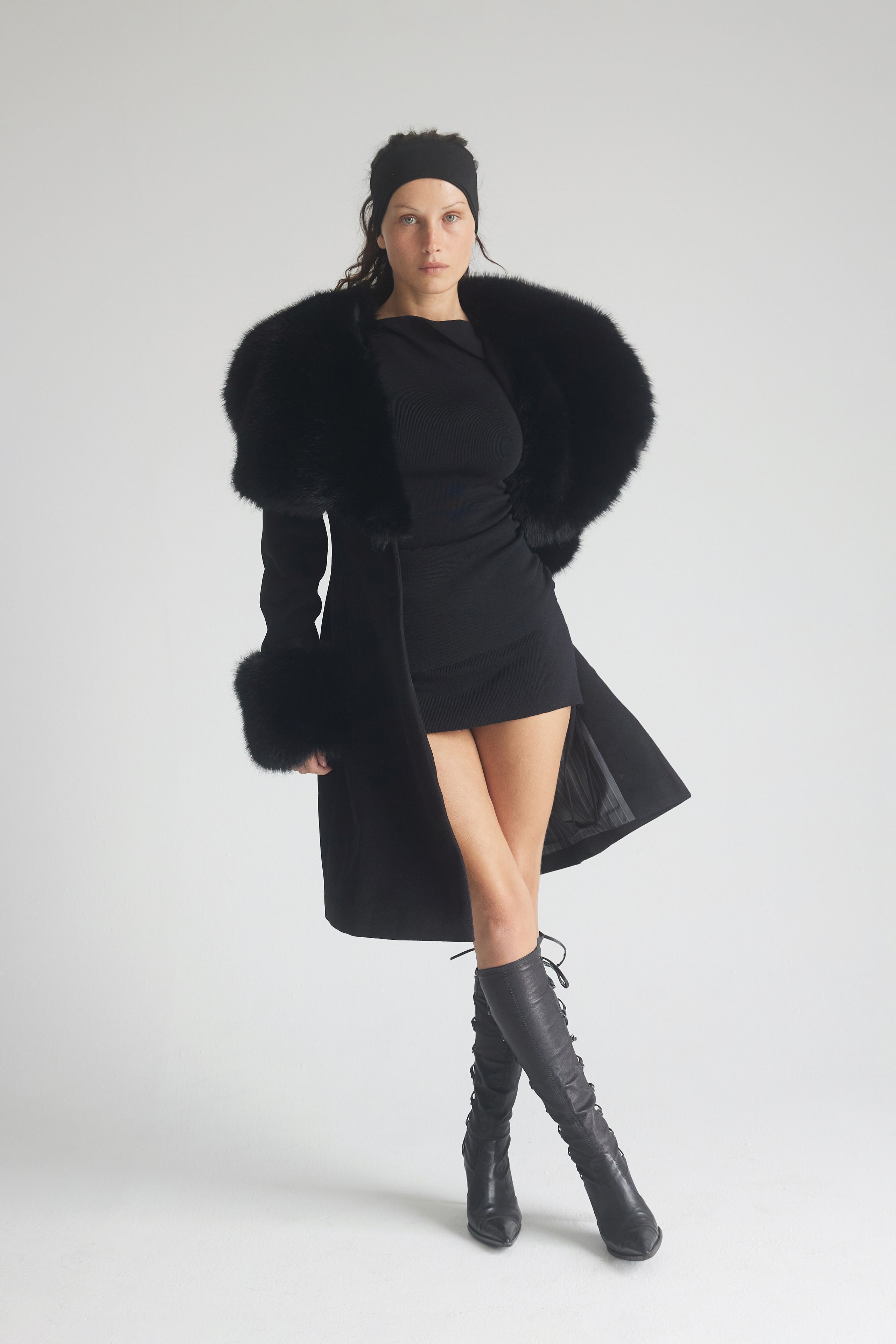 Woman wearing a black coat with fur trim, black dress, and black boots on a white background